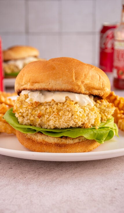 A crispy baked fish fillet sandwich on a soft bun with tartar sauce and lettuce on a white plate with french fries.