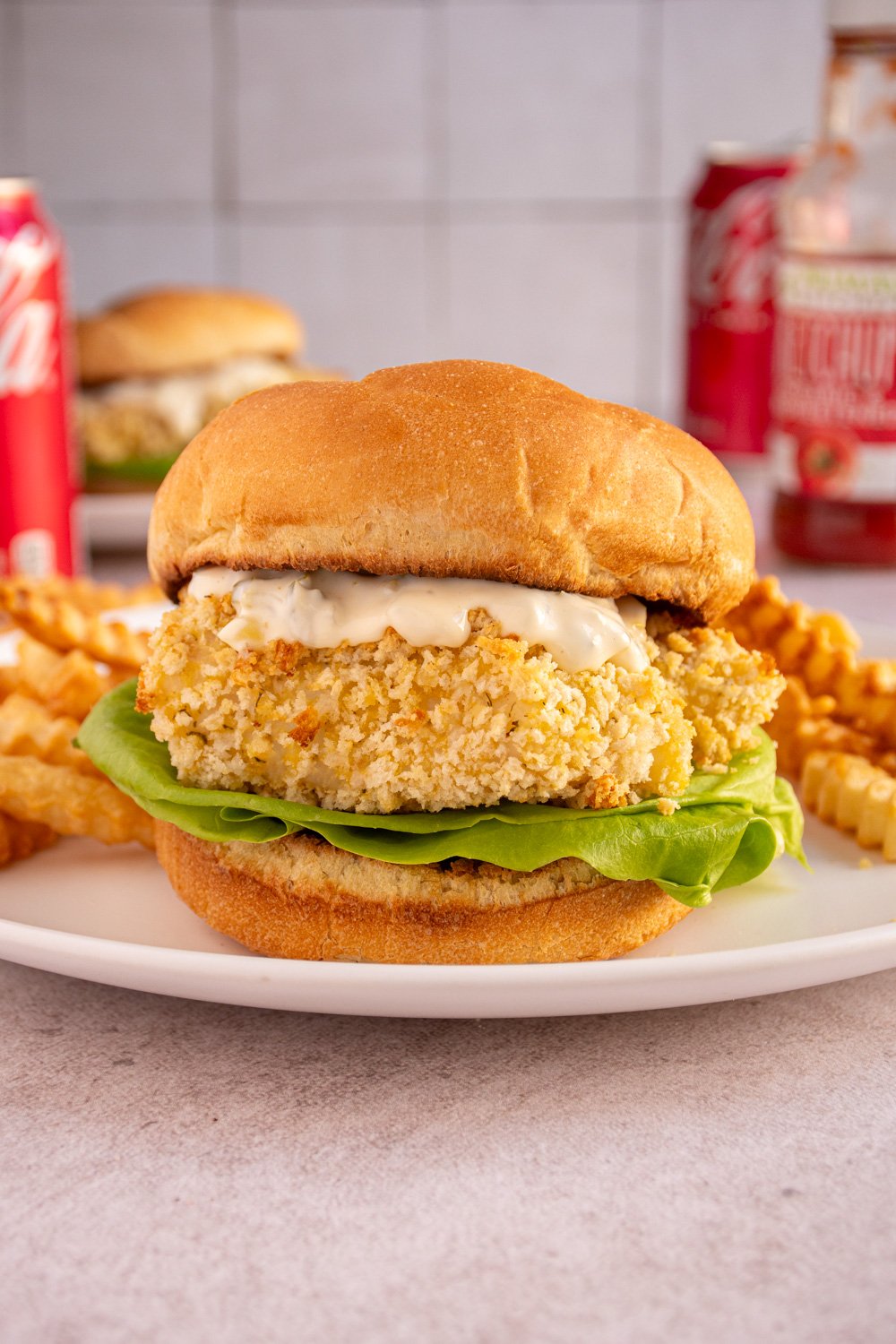 A crispy baked fish fillet sandwich on a soft bun with tartar sauce and lettuce on a white plate with french fries.