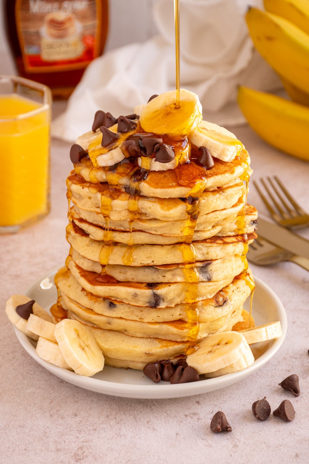 A large stack of Banana Chocolate Chip Pancakes topped with chocolate chips and slices of bananas with syrup drizzling from the top down the sides.