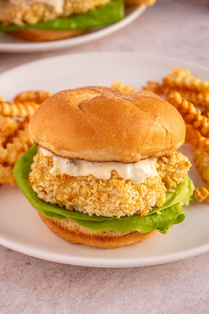 A baked fish sandwich between to soft buns with tartar sauce and lettuce on a white plate next to french fries.