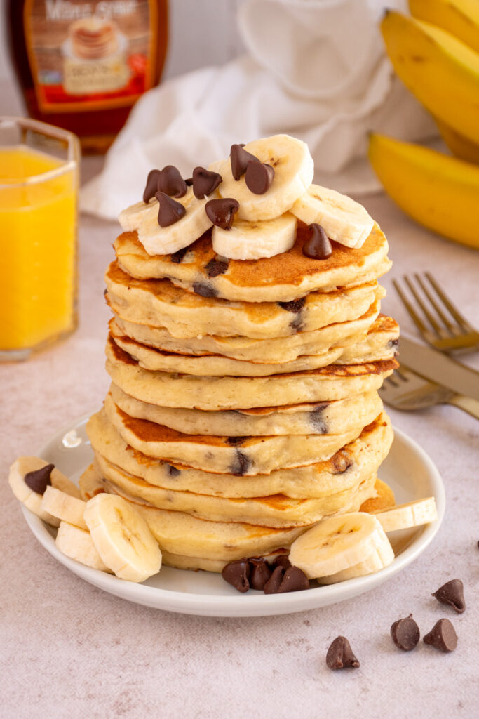 A plate with a large stack of Banana Chocolate Chip Pancakes topped with chocolate chips and banana slices.