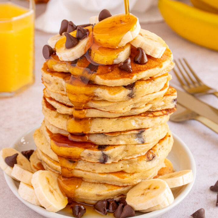 Maple syrup being poured on to stack of Banana Chocolate Chip Pancakes topped with chocolate chips and banana slices.