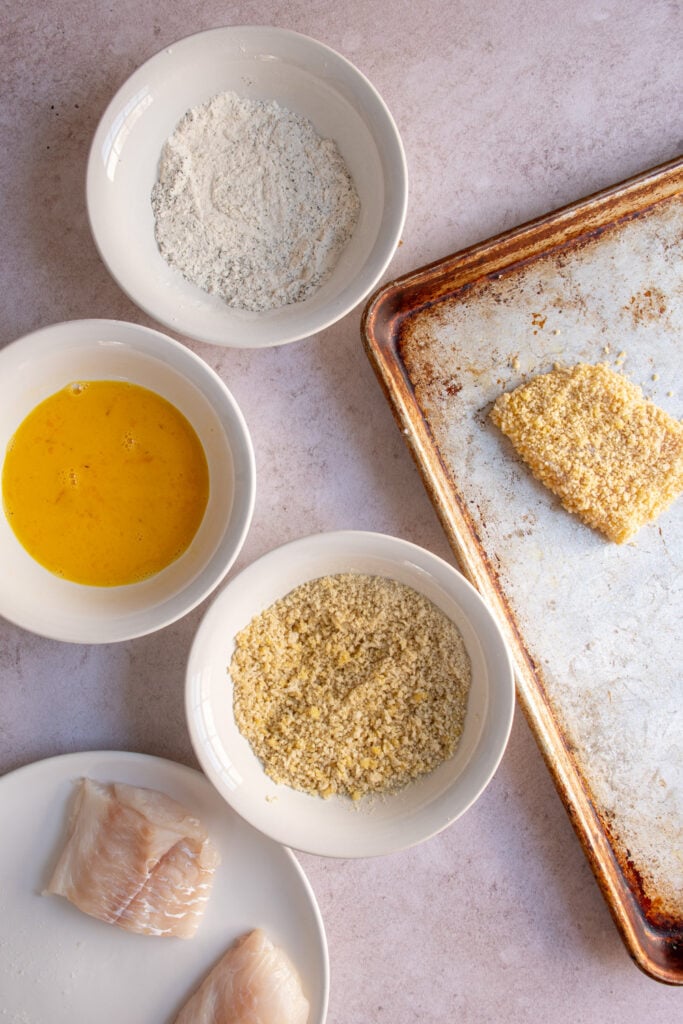 The set up with bowls for a flour mixture, egg, and breadcrumbs to coat and bread a piece of fish.