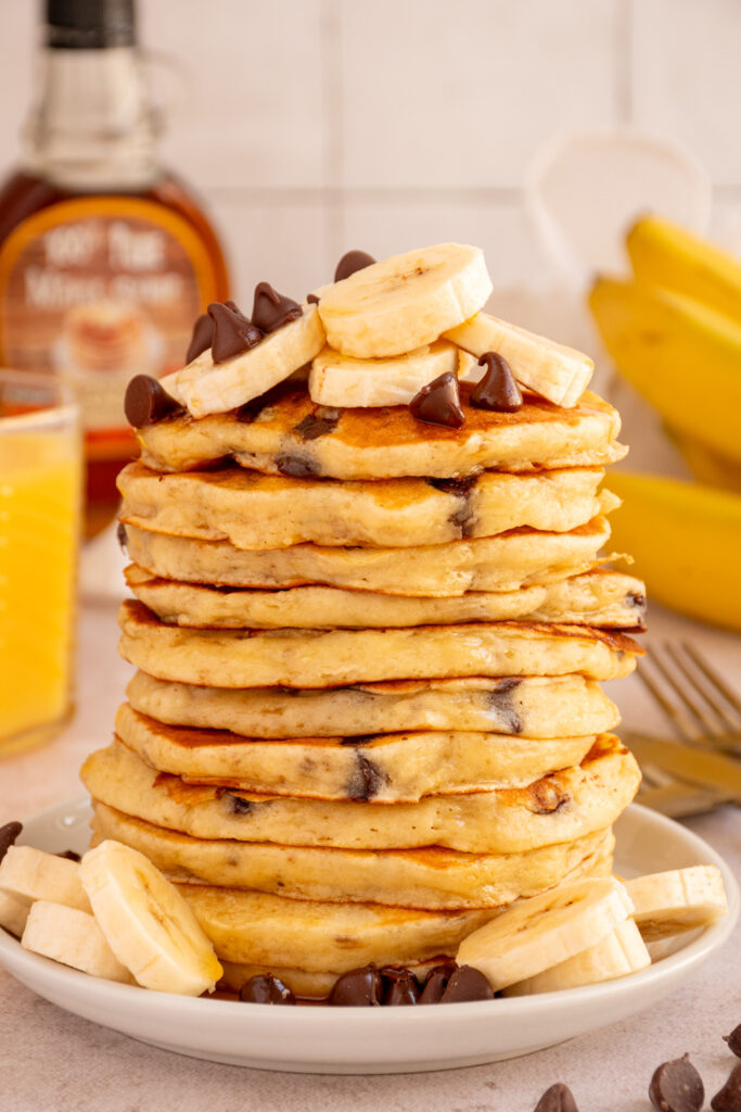 A very large stack of Banana Chocolate Chip Pancakes on a plate topped with banana slices and chocolate chips.