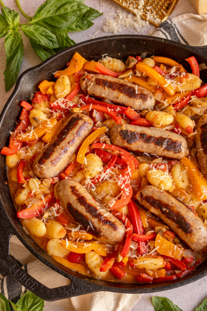 A close up of a large skillet with colorful bell peppers, gnocchi, and browned sausages.