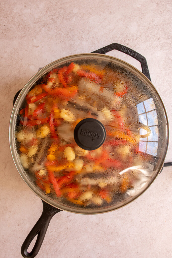A covered skillet with peppers and sausages inside.
