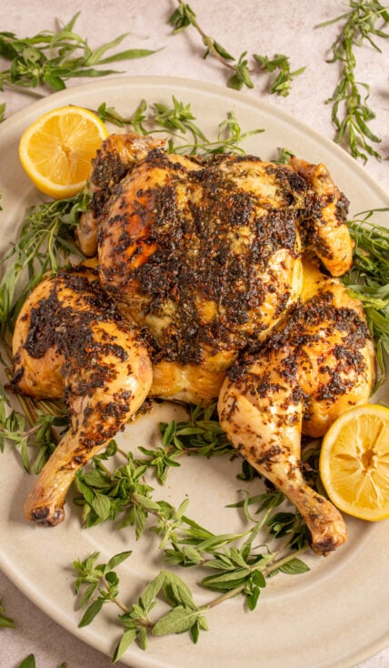A butterflied roasted chicken on a white serving platter surrounded by green fresh herbs.