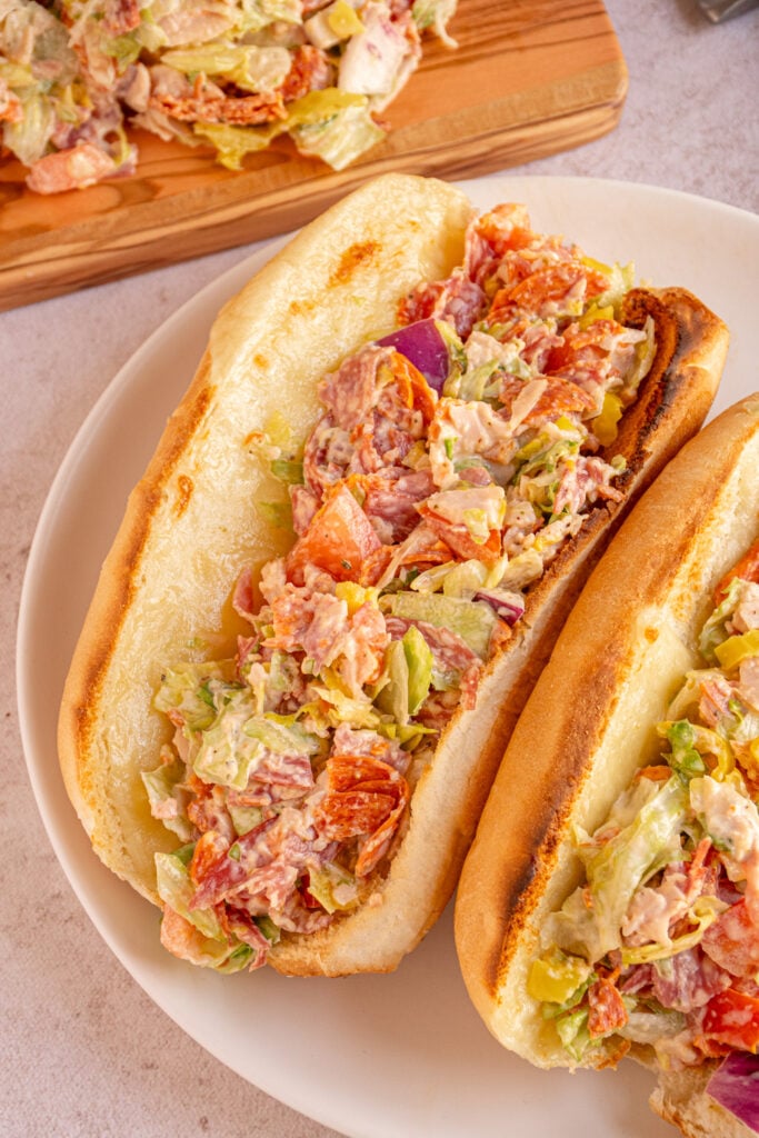 A pair of Chopped Italian Sandwiches on a white plate sitting next to a cutting board with chopped fillings.