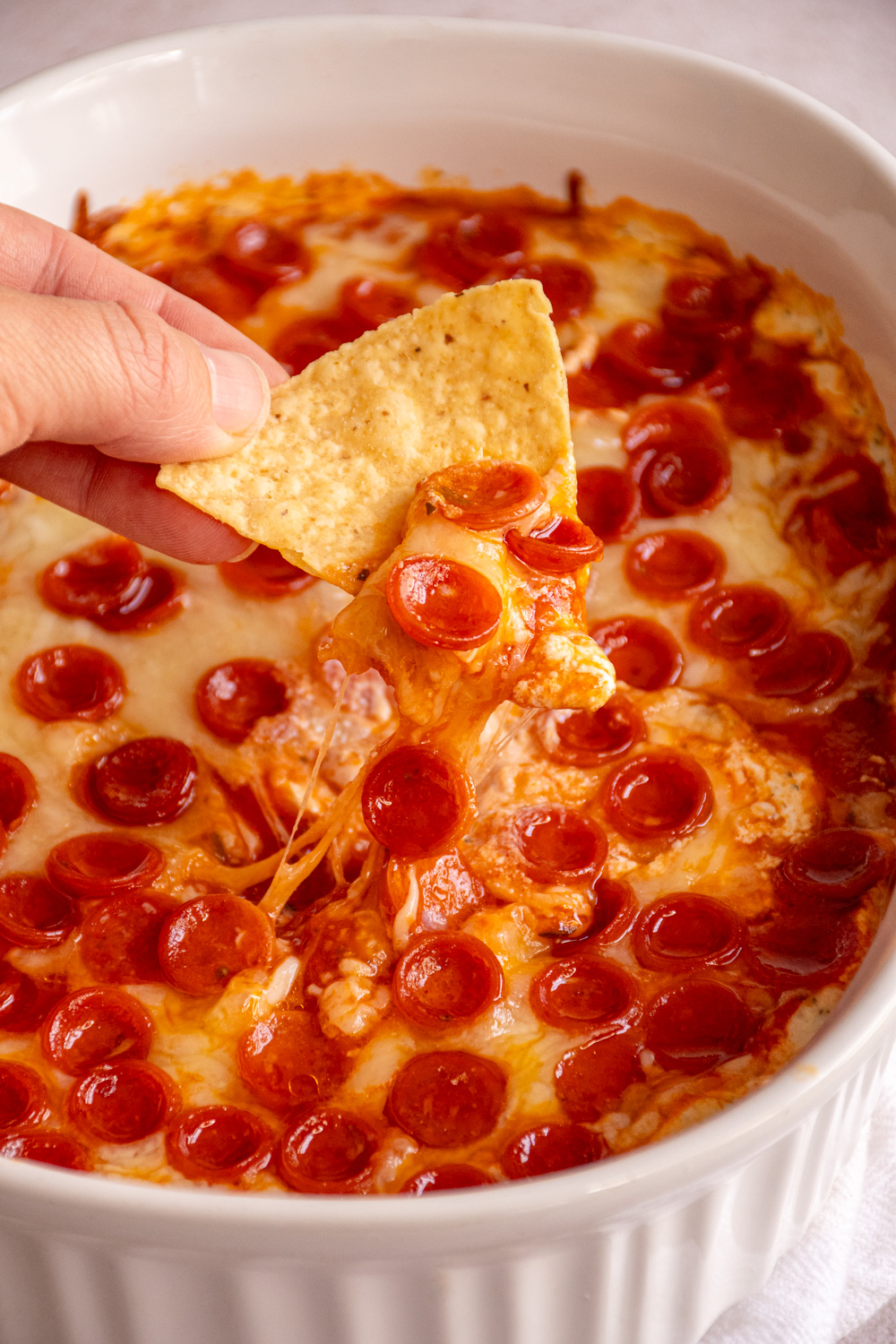 A casserole dish filled with cottage cheese pizza dip covered in mini pepperoni with a tortilla chip being dipped and pulling along melted cheese.