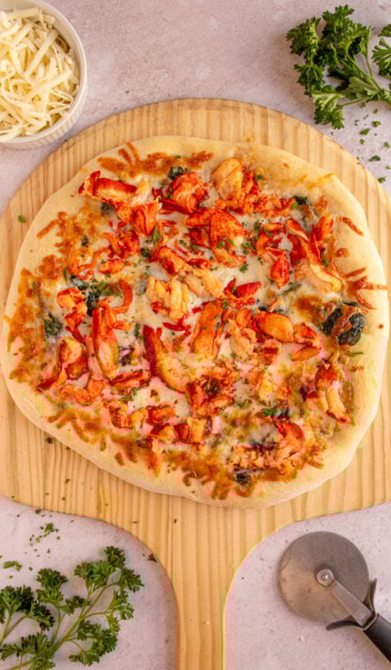 A large lobster pizza on a wooden pizza peel surrounded by fresh parsley.