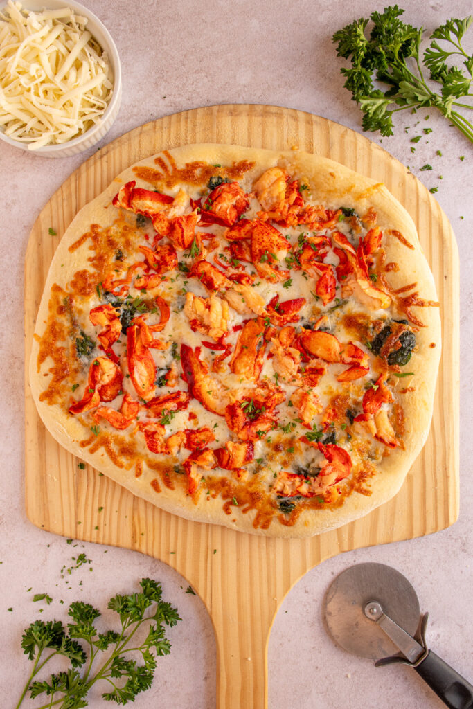 A large lobster pizza on a wooden pizza peel surrounded by fresh parsley.