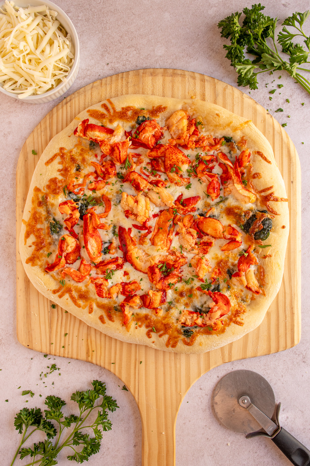 A large lobster pizza on a wooden pizza peel surrounded by fresh parsley.
