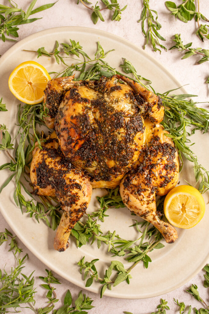 A serving platter holding a spatchcocked chicken crusted with fresh herbs and surrounded by fresh herbs.
