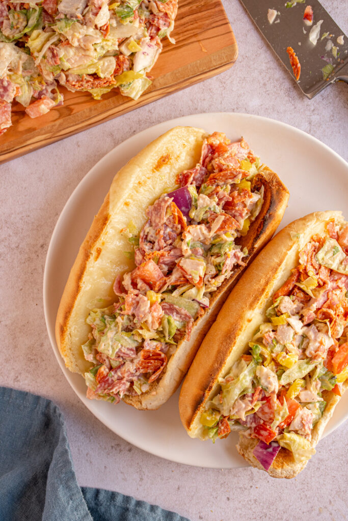 A white plate with two Chopped Italian Sandwiches laying on it filled with meats, veggies, and cheese.