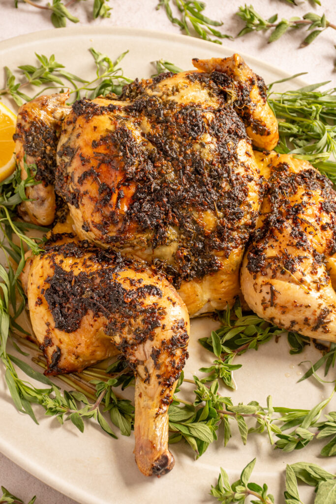 A butterflied roast chicken covered in baked herbs on a serving platter surrounded by fresh herbs.