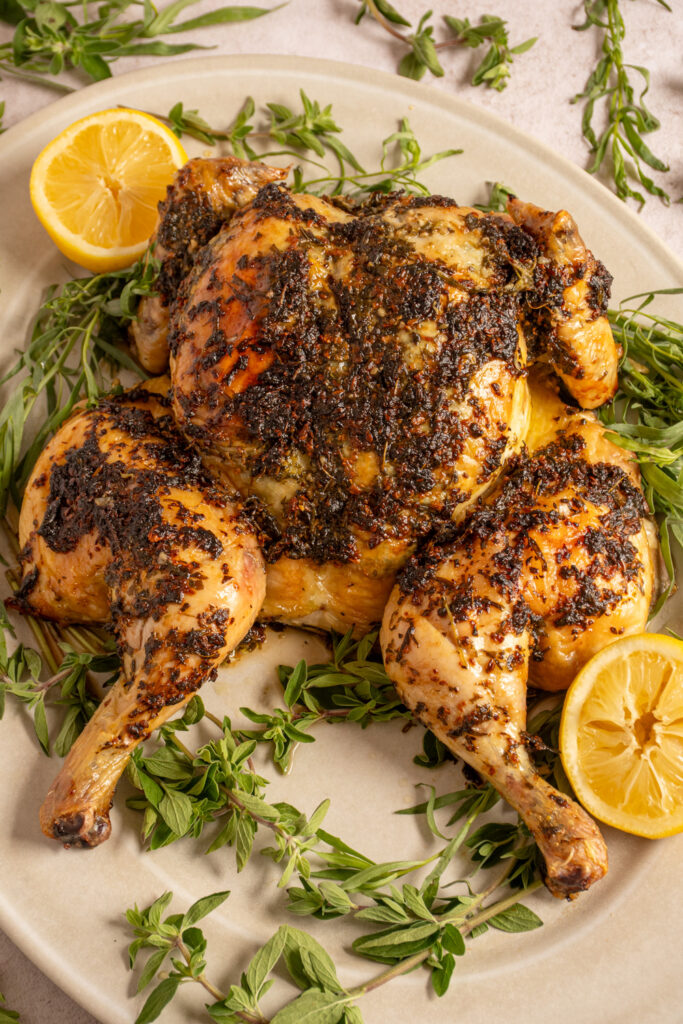 Herb crusted butterflied roast chicken on a serving platter surrounded by fresh tarragon and oregano.