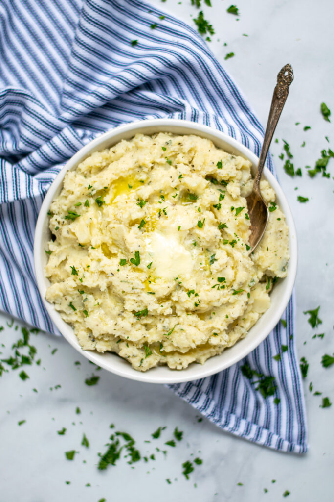 A large white bowl filled with herbed mashed potatoes and covered in melted butter with a spoon for serving. 