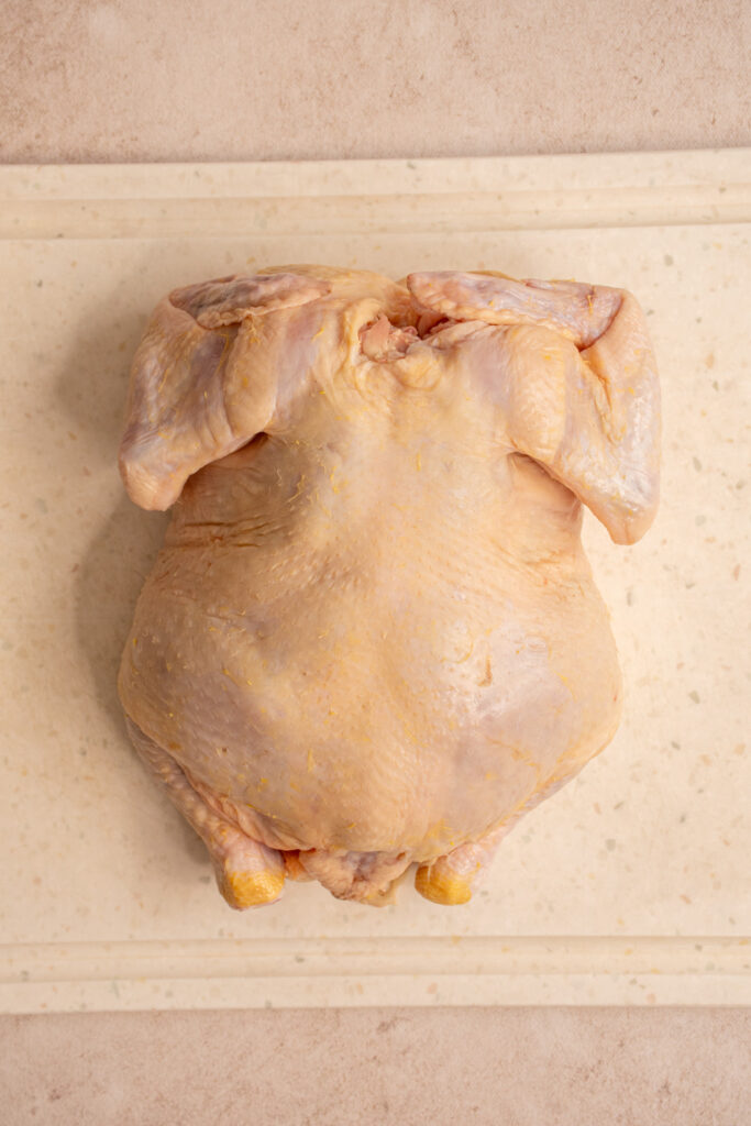 A whole raw chicken breast side down on a cutting board showing the backbone.