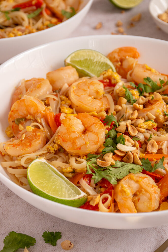 A white bowl filled with shrimp pad thai filled with noodles, shrimp and veggies garnished with cilantro, peanuts, and lime wedges.