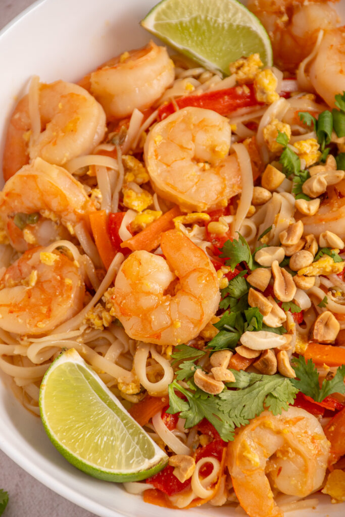 Close up of shrimp pad thai in a bowl with rice noodles, veggies, and garnished with cilantro, lime wedges, and chopped peanuts.