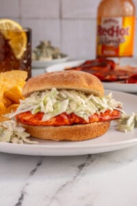 A grilled buffalo chicken sandwich with blue cheese coleslaw on a white plate next to some chips.