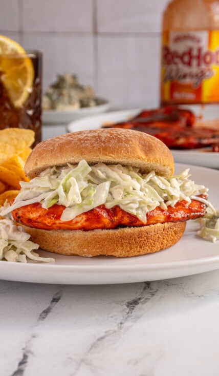 A grilled buffalo chicken sandwich with blue cheese coleslaw on a white plate next to some chips.