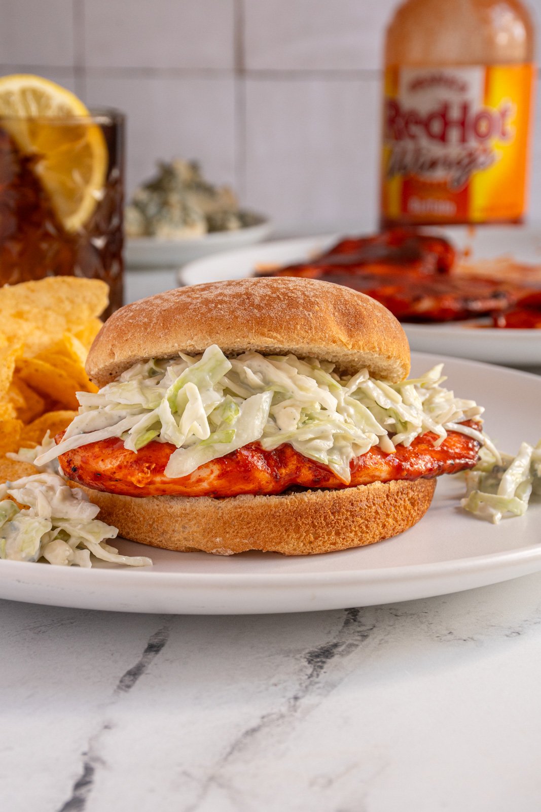 A grilled buffalo chicken sandwich with blue cheese coleslaw on a white plate next to some chips.