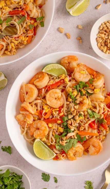 A large white bowl filled with shrimp pad thai garnished with lime wedges, cilantro, and chopped peanuts.