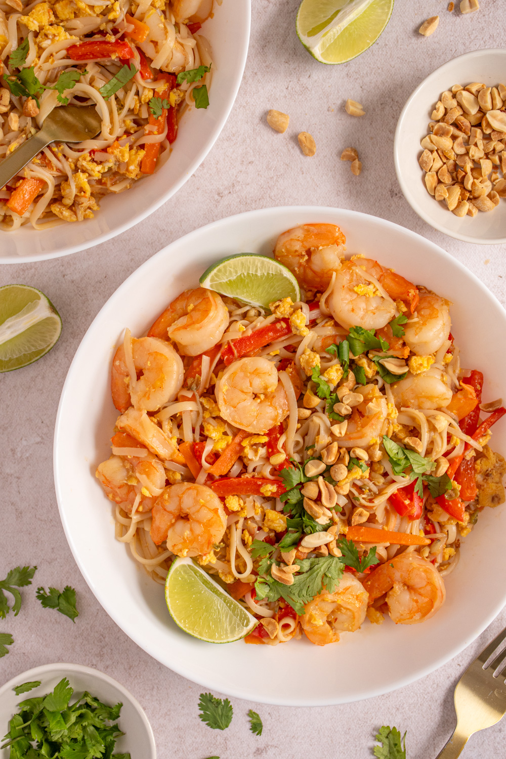 A large white bowl filled with shrimp pad thai garnished with lime wedges, cilantro, and chopped peanuts.
