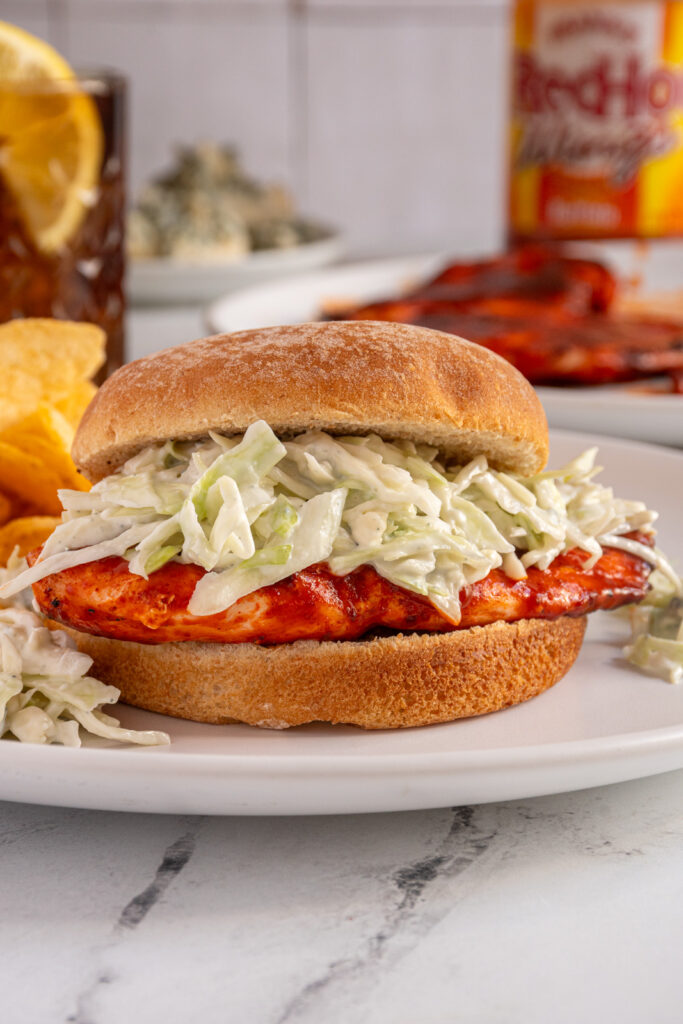 A close up of a grilled buffalo chicken sandwich with buffalo chicken topped with blue cheese coleslaw on a white plate.