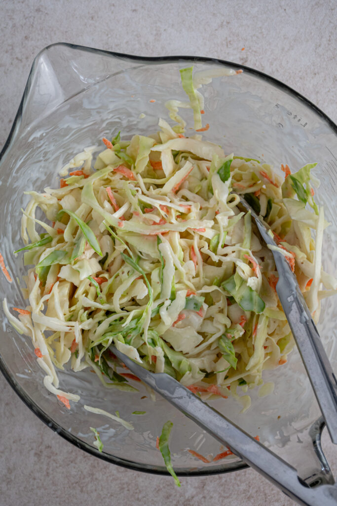 A Mixing bowl and tongs mixing together coleslaw dressing and coleslaw.