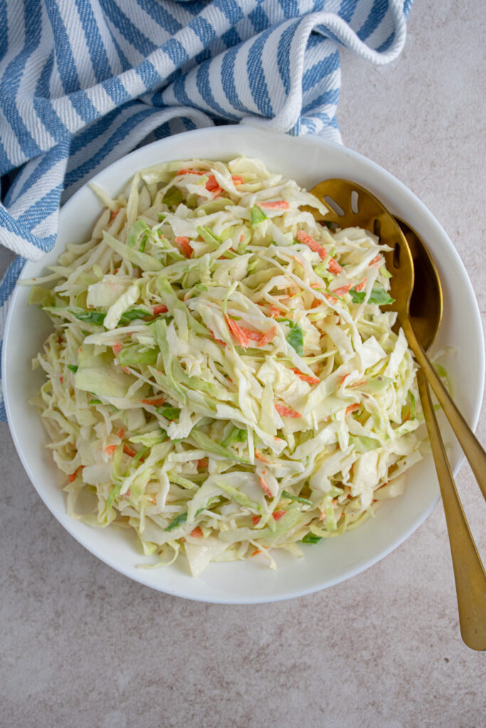 Overhead view of a white serving bowl filled with coleslaw recipe without celery seed and gold serving spoons.