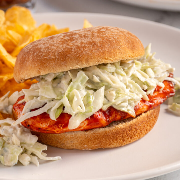 A white plate with a grilled buffalo chicken sandwich topped with blue cheese coleslaw.