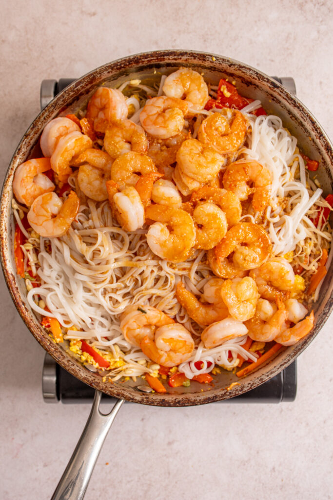 Adding the noodles, shrimp, and sauce to the vegetables in a skillet.