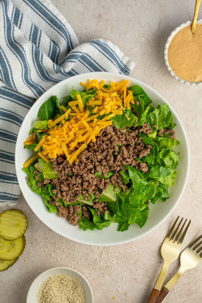 A white salad bowl filled with chopped romaine lettuce, browned ground beef, and shredded cheddar cheese.