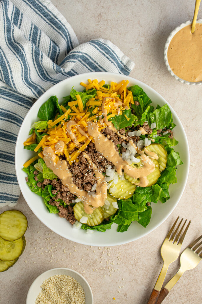 A white salad bowl filled with chopped romaine lettuce, browned ground beef, shredded cheddar cheese. pickle chips, diced sweet onion, special sauce, and sesame seeds.