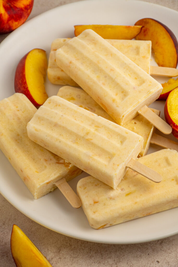 Creamy peach yogurt popsicles on a white plate with slices of fresh peach.