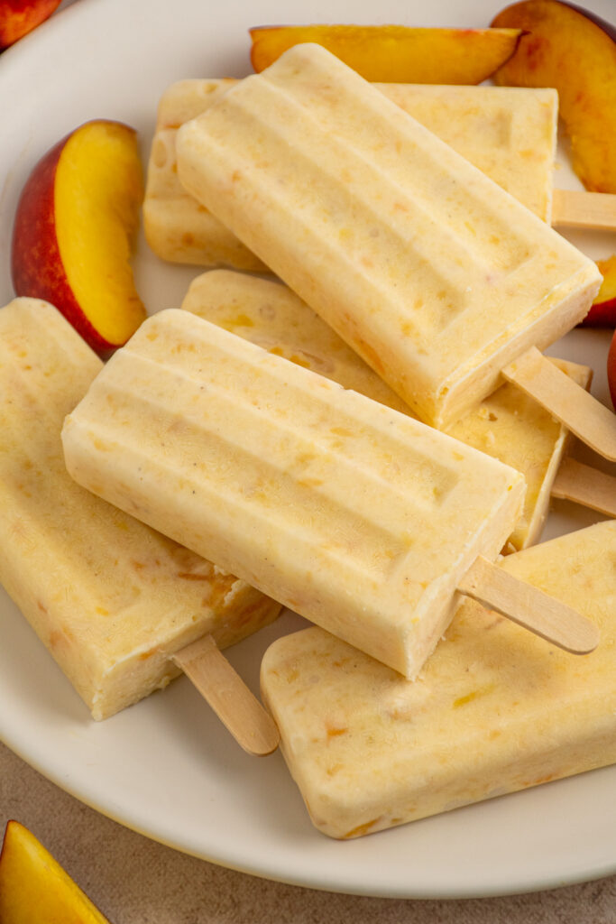 A pile of peach yogurt popsicles on a white plate surrounded by fresh peach slices.
