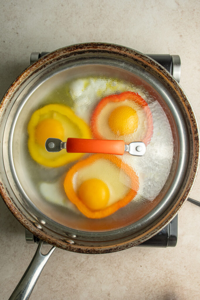 A covered skillet with cooking eggs in pepper rings.