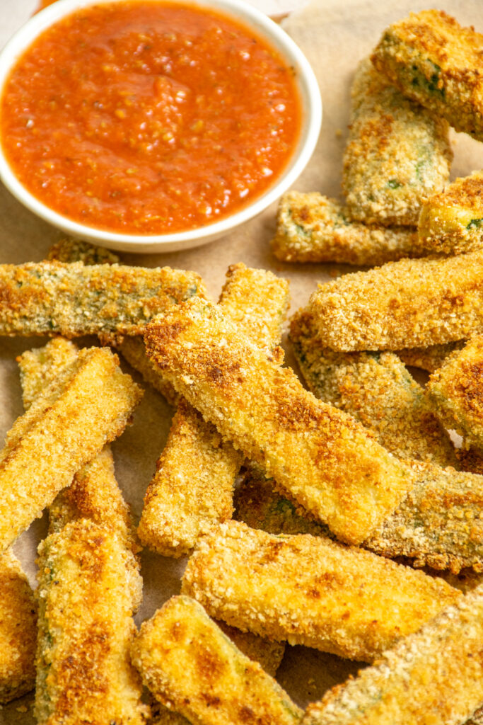 Baked zucchini fries on a platter sitting next to a cup of marinara sauce.