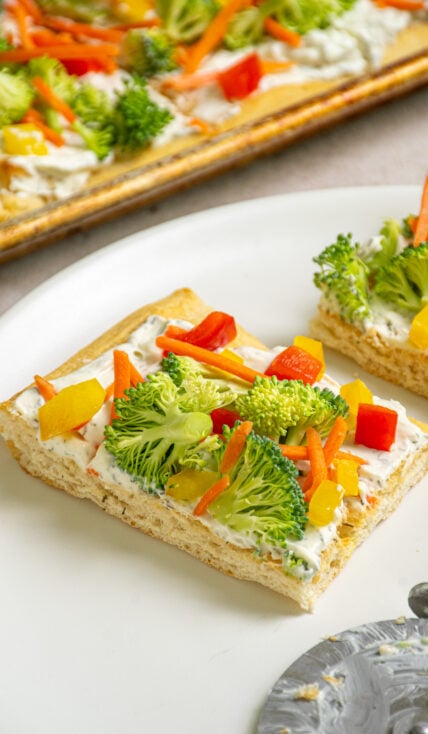 A slce of crescent roll veggie pizza on a white plate topped with fresh broccoli, carrots, and bell peppers.