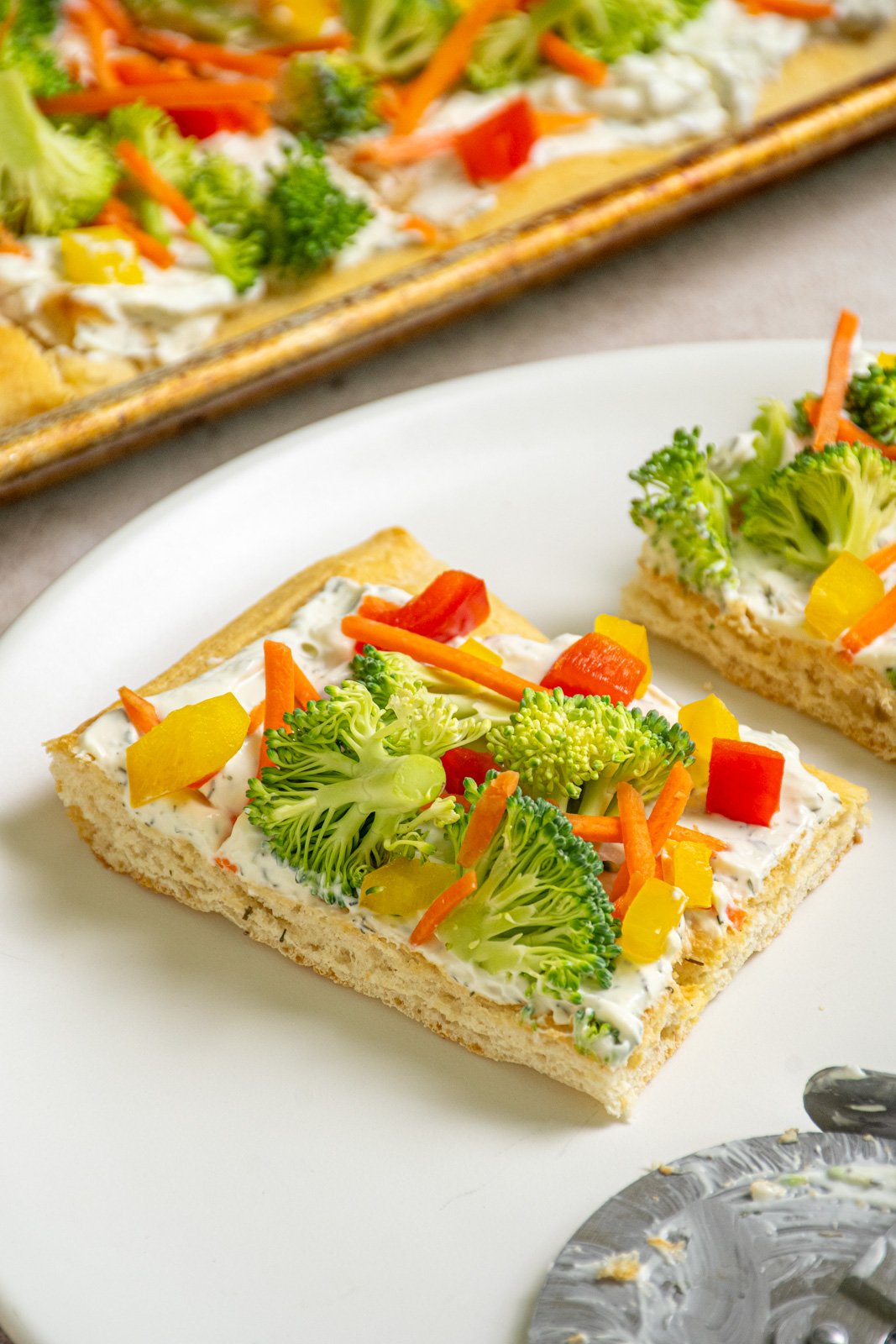 A slce of crescent roll veggie pizza on a white plate topped with fresh broccoli, carrots, and bell peppers.