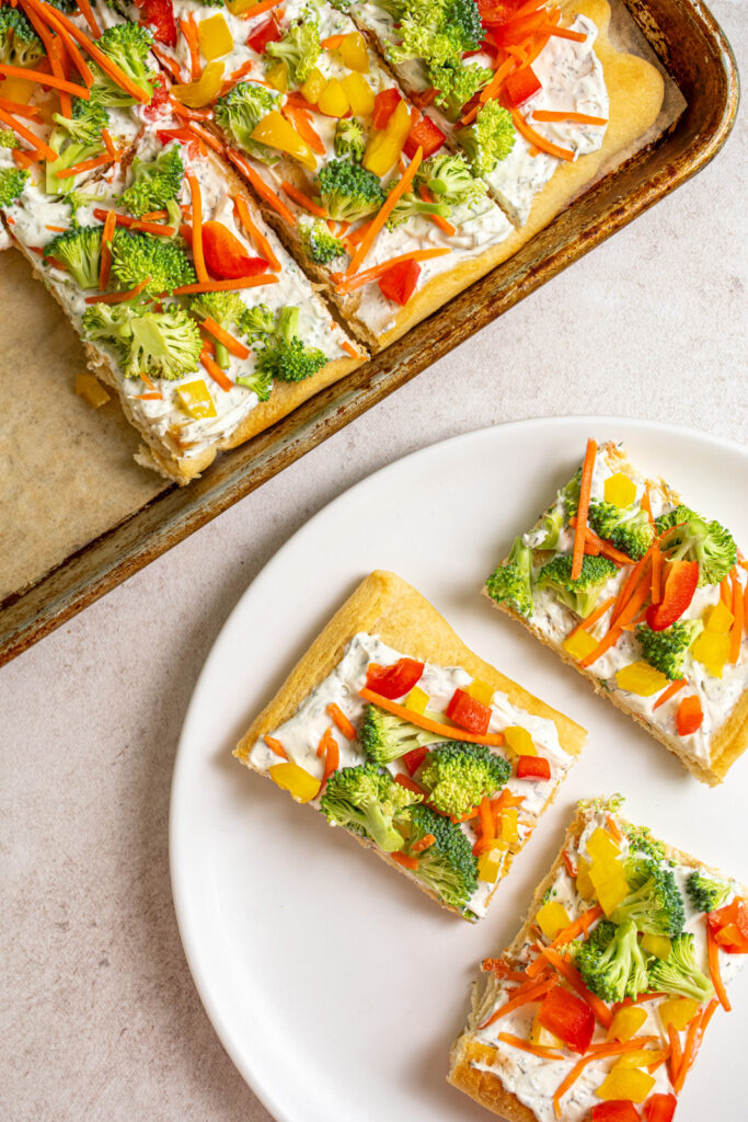 Overhead view of the sheet pan and white plate with slices of cold veggies pizza on them.