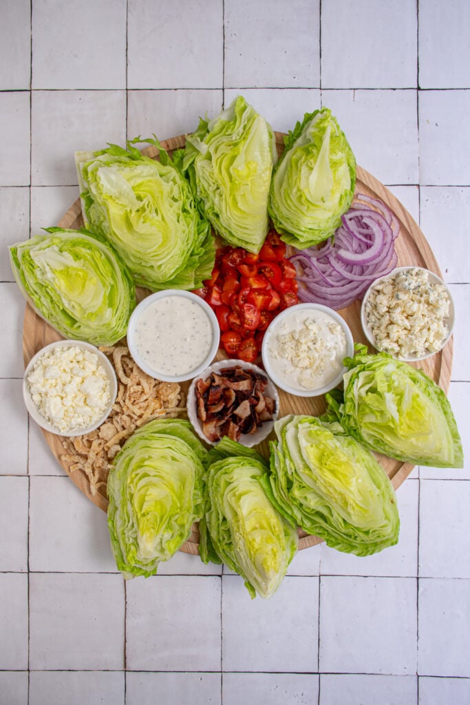 Overhead view of a large wooden board filled with makings for wedge salad like iceberg lettuce, dressings, and toppings.