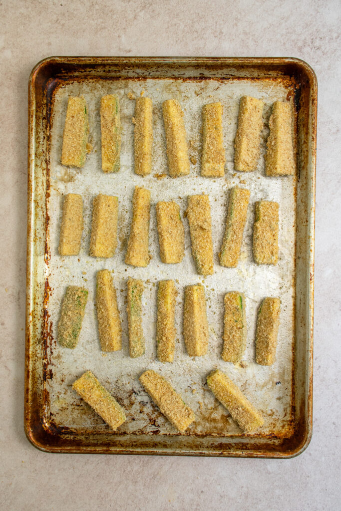 Uncooked breaded zucchini fries on a greased sheet pan.