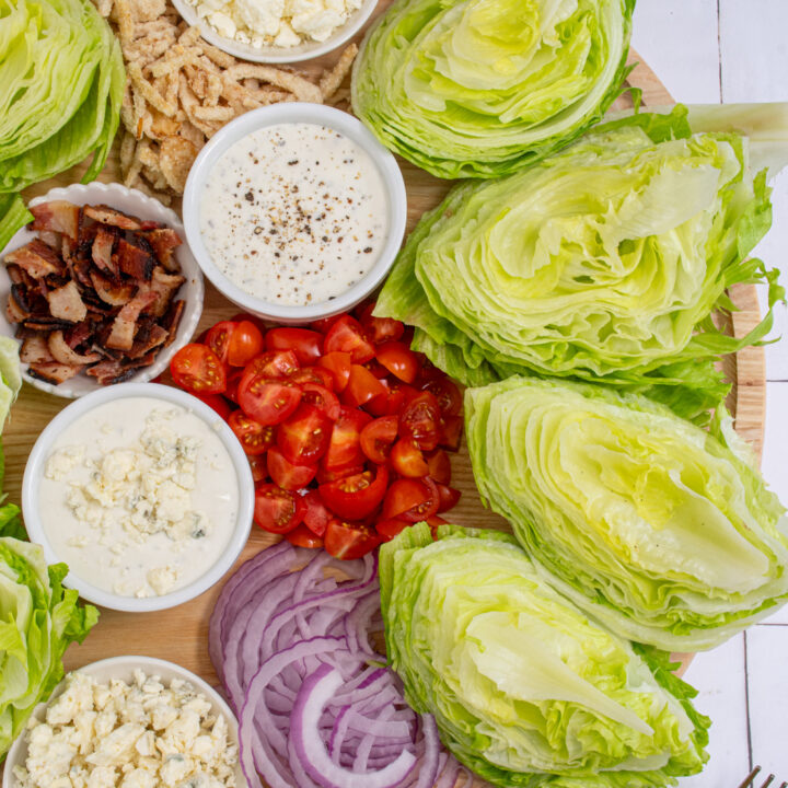 Large circle wooden board filled with makings of a classic wedge salad like iceberg lettuce, dressings, and toppings.