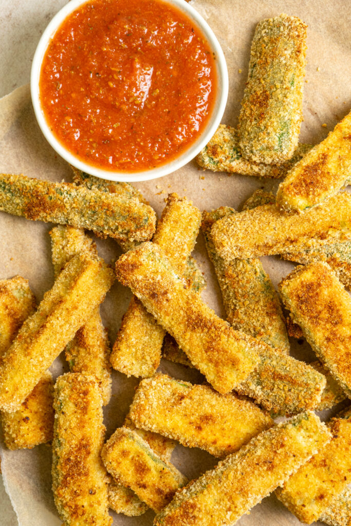 Oven Baked Crispy Zucchini Fries on brown parchment paper with a side of marinara sauce.