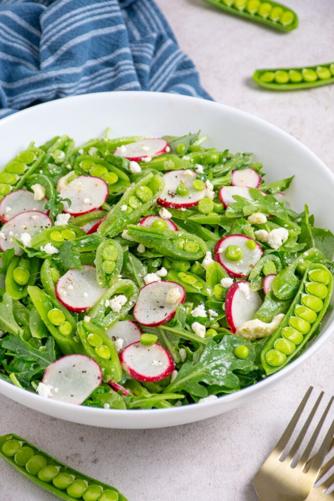 A white bowl filled with sugar snap pea salad with arugula, radishes, and feta.