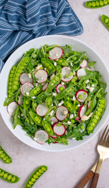 A white bowl filled with sugar snap pea salad with arugula, radishes, and feta.