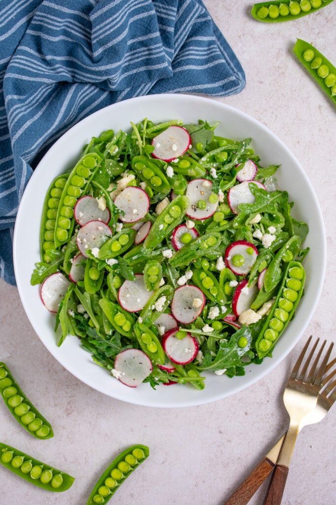 A white bowl filled with sugar snap pea salad with arugula, radishes, and feta.